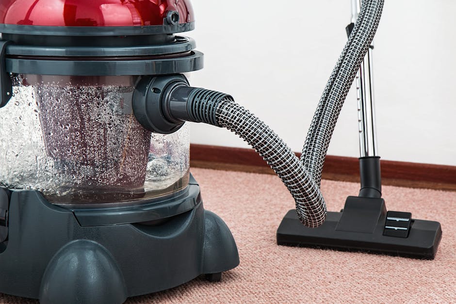 A person using a vacuum cleaner on a patterned, beige and green floral area rug in a room. The individual is wearing beige shoes and blue jeans, with a beige jacket, and is holding the vacuum's handle