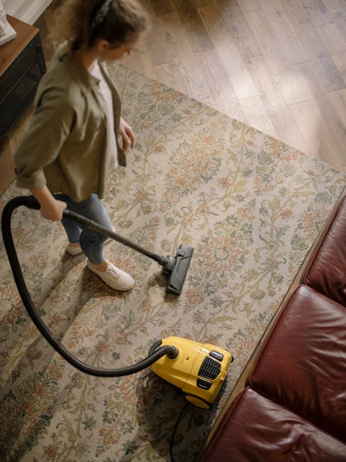 A woman is engaged in surface cleaning using a yellow vacuum cleaner on a large, traditional patterned rug in a living room. The room features hardwood flooring adjacent to the rug and contains a leat