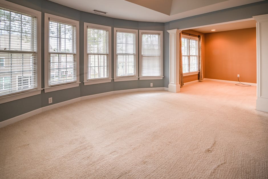 A spacious residential living room featuring a light beige carpeted floor that appears clean and plush, with a few subtle indentations visible. The room has a curved wall lined with six large double-h